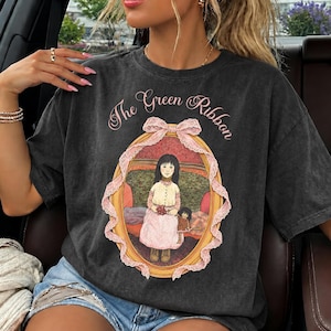以下が含まれることがあります： チャコールグレーの半袖Tシャツ。ピンクのドレスを着て人形を持つ少女のグラフィックプリントが施され、ピンクのレースリボンが付いた金色の楕円形の枠で囲まれています。枠の上部には筆記体で「The Green Ribbon」という文字が印刷されています。