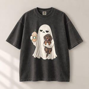 May include: A charcoal grey short-sleeve t-shirt featuring a graphic of a white ghost holding a tan iced coffee drink and carrying a small brown and black dachshund puppy. The ghost has a simple face with two black eyes and a straight mouth line.
