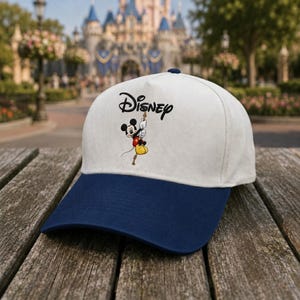May include: A baseball cap with a white crown and a dark blue brim. The front features an embroidered black Disney logo above a graphic of Mickey Mouse in a red shirt and yellow shorts, shown hanging from the letter y in the Disney text.