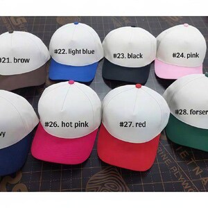 May include: Eight baseball caps with white crowns and colored brims, labeled #21 brown, #22 light blue, #23 black, #24 pink, #25 navy, #26 hot pink, #27 red, and #28 forest green. Each cap features a small button on the top center of the crown.