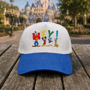 May include: A white baseball cap with a blue brim featuring the word "HEY!" in colorful letters. The letters are held up by cartoon characters including Goofy, Mickey Mouse, Donald Duck, and Pluto. The background shows a castle.