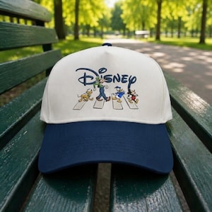 May include: A white and navy blue baseball cap featuring the Disney logo and characters crossing a street. The cap has a curved brim and is embroidered with a colorful design of Goofy, Donald Duck, Mickey Mouse, and Pluto.