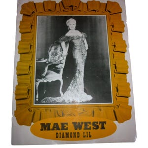 May include: A black and white photograph of Mae West in a glamorous gown, framed in a gold-colored border with a decorative ribbon design. The text "MAE WEST DIAMOND LIL" is displayed in a gold oval at the bottom.