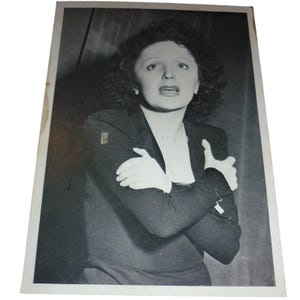 May include: Black and white photograph of a woman with dark, curly hair, wearing a dark blazer and white gloves. She has her arms crossed and is looking up with an open mouth. The image is on a rectangular piece of paper.