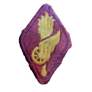 May include: A diamond-shaped embroidered patch featuring a deep purple background with a central yellow design. The design depicts a vertical torch combined with a winged wheel, rendered in yellow thread against the purple fabric base.