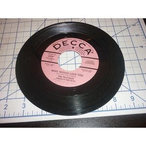May include: A black 45 RPM vinyl record with a pink Decca label featuring the text Who Would Love You by The Tell Tales. The record is positioned on a white cutting mat with a blue grid, showing a diameter of approximately 7 inches.
