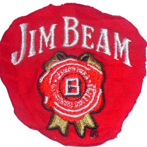 May include: A circular red fabric patch featuring the white embroidered text Jim Beam above a central red wax seal graphic. The seal contains a white letter B, surrounded by gold ribbon accents and black outlines, creating a detailed emblem.
