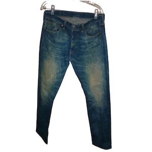 Könnte beinhalten: Blaue Jeans aus Denim mit Knopfleiste und klassischem Fünf-Taschen-Design. Die Jeans haben eine verwaschene Waschung mit helleren Bereichen an Oberschenkeln und Knien. Sie hängen an einem durchsichtigen Kunststoffbügel.