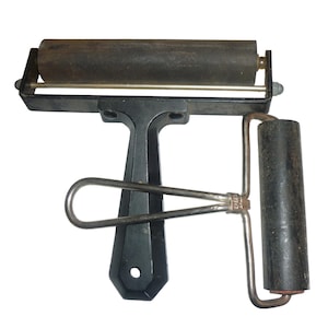 Könnte beinhalten: Ein Vintage-Druckwalze mit einer dunkelgrauen Walze und einem schwarzen Griff. Die Walze hat einen Metallrahmen und eine kleinere Walze am Griff. Das Werkzeug dient dazu, Tinte gleichmäßig auf eine Oberfläche aufzutragen.