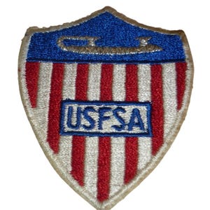 May include: A shield-shaped patch with a blue top section and red and white stripes below. The top has a silver design and the text "USFSA" is in a blue rectangle.