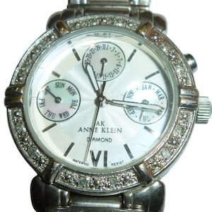 Puede incluir: Reloj Anne Klein plateado con esfera blanca y diamantes en el bisel. Muestra el día, la fecha y el mes, con números romanos y el texto "WATER 100 RESIST".
