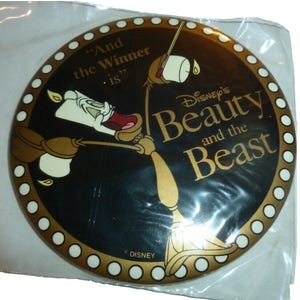 May include: A round button featuring characters from Disney's Beauty and the Beast. The button has a black background with gold accents and the text "And the Winner is." The Disney logo is at the bottom.