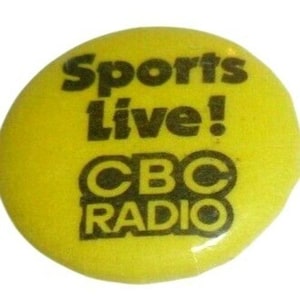 May include: A yellow button featuring the text "Sports Live!" in black, above "CBC RADIO" in black. The button is circular and has a shiny surface. The image is a close-up of the button.