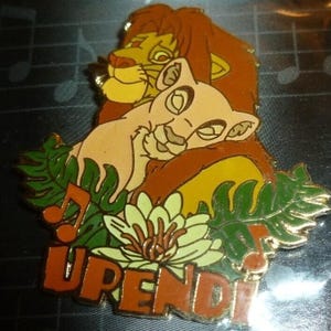 May include: A colorful enamel pin featuring Simba and Nala from The Lion King. The pin shows Simba embracing Nala, with the word "UPENDI" in orange letters at the bottom. Green leaves and a flower are also included in the design.