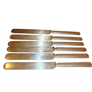 Può includere: Un set di sette coltelli da burro vintage in argento. Ogni coltello ha una lama rettangolare e un manico con un design a conchiglia decorativo. I coltelli sono disposti su uno sfondo bianco.