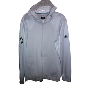May include: Light gray Adidas zip-up hoodie with a hood and front pockets. Features the Adidas logo on the right sleeve and a black and white logo on the left sleeve. The jacket has a full-length zipper and a gray lining.