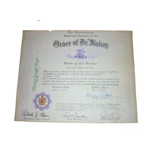 Może przedstawiać: Zabytkowy papierowy certyfikat Międzynarodowej Najwyższej Rady Zakonu DeMolay z 1974 roku. Dokument zawiera czarny drukowany tekst, fioletowy emblemat oraz odręczne podpisy wykonane niebieskim i czarnym tuszem na złamanej bieli tła.