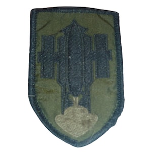 Pode incluir: Um emblema bordado em forma de escudo com um fundo verde-oliva e um emblema central azul-marinho escuro representando uma forma vertical de foguete ou míssil ladeada por dois retângulos, com uma forma de nuvem bege clara na base.