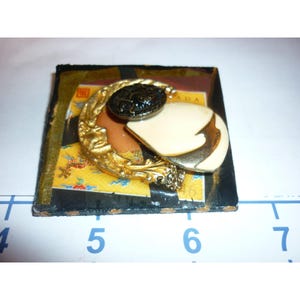 Puede incluir: Broche vintage con diseño de media luna dorada, botón negro y forma crema. El broche está sobre una base cuadrada con un fondo estampado amarillo y negro. El broche mide aproximadamente 5 cm de largo.