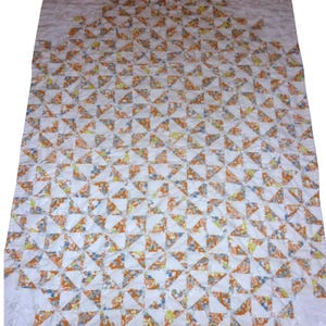 May include: A rectangular quilt featuring a repeating pattern of white triangles and floral-printed triangles in shades of orange, yellow, and blue. The fabric pieces are stitched together in a grid, creating a geometric design across the surface.