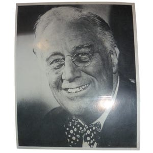 Könnte beinhalten: Schwarz-Weiß-Fotografie eines lächelnden Mannes mit Brille und einer gepunkteten Fliege. Der Mann hat kurze, graue Haare und trägt einen Anzug. Das Foto hat einen weißen Rahmen.