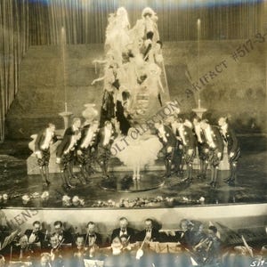 May include: A black and white vintage photograph depicting a theatrical stage performance. A central figure in a white tutu stands on a circular platform surrounded by performers in dark tailcoats and breeches, with an orchestra in the foreground.
