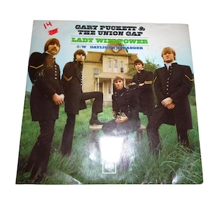 May include: Album cover for "Lady Willpower" by Gary Puckett & The Union Gap. The cover features a photograph of the band members in black military-style jackets and pants, standing in front of a house. The album title and band name are printed in green and black.