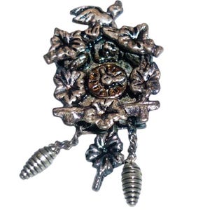 May include: A silver-toned cuckoo clock brooch with intricate floral and bird details. The brooch features a central clock face, decorative leaves, and hanging pendulum weights. The design is detailed and vintage-inspired.