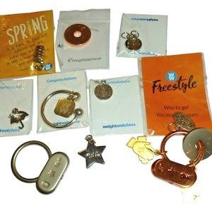 Op de afbeelding: Een verzameling Weight Watchers mijlpaalbedels en sleutelhangers, inclusief diverse metalen vormen zoals een ster, paraplu en ronde penningen. Sommige items zijn bevestigd aan merkkaarten met tekst zoals Spring, Congratulations en Freestyle.