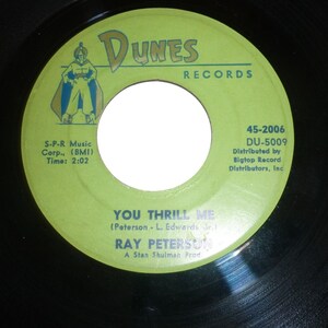 May include: A vintage 45 RPM vinyl record with a lime green label. The label features the text "DUNES RECORDS" and the song title "YOU THRILL ME" by Ray Peterson. The record has a white center and black outer edge.