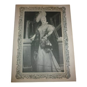May include: Black and white vintage print of a woman in a long dress, hat, and fur stole. The image is framed with a decorative border. The woman is standing near a railing, holding a small purse.