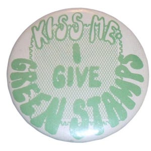May include: A round pinback button featuring a white background with light green text that reads KISS ME I GIVE GREEN STAMPS. The design includes a light green grid pattern behind the central text, creating a distinct visual contrast on the surface.