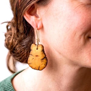 May include: A drop earring featuring a light brown, irregularly shaped wooden pendant with visible dark grain lines and a smooth finish. The pendant is suspended from a thin, gold-toned metal wire hook that passes through the top of the wood piece.