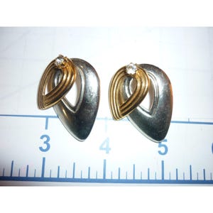May include: A pair of vintage earrings with a silver-tone teardrop shape and a gold-tone accent. Each earring features a small, clear gemstone at the top. The earrings are displayed on a white surface.