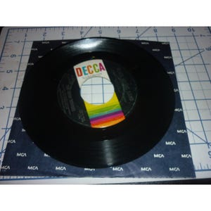 May include: A black vinyl 45 RPM record featuring a Decca label with a rainbow-colored stripe design. The record is placed on a dark blue paper sleeve with a repeating white MCA logo pattern, set against a white grid cutting mat background.