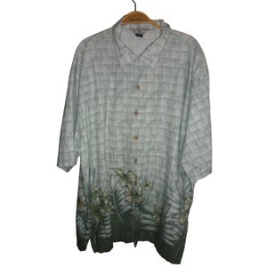 May include: A light green button-down shirt with a square pattern and floral design. The shirt has short sleeves and a collar. The bottom of the shirt features a dark green band with white and green flowers and leaves.