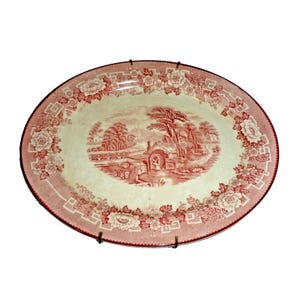 May include: An oval ceramic platter featuring a red and white transferware pattern. The center depicts a rural landscape with a water mill and trees, while the border is decorated with a repeating floral motif and geometric Greek key accents.