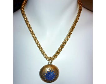 Puede incluir: Collar dorado con cadena de trigo y colgante redondo. El colgante presenta un diseño floral azul en filigrana dorada. El collar se exhibe en un maniquí blanco.
