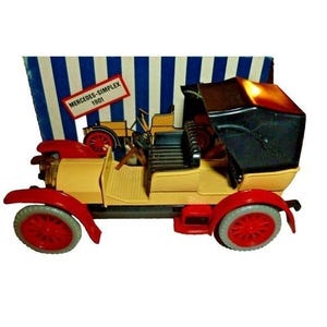Peut inclure: Une voiture jouet de couleur crème, avec des ailes rouges et des roues rouges. La voiture a un toit décapotable noir et une petite enseigne avec l'inscription "MERCEDES-SIMPLEX 1901". Le fond présente des rayures bleues et blanches.