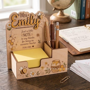 Può includere: Organizzatore da scrivania in legno con un design personalizzato per un insegnante. Presenta un porta blocco note, fessure per penne ed elementi decorativi come api, fiori e un vasetto di miele. Il testo recita "Mrs. Emily" e un messaggio di ringraziamento.