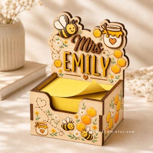 Può includere: Organizzatore da scrivania in legno a tema api e favi. L'organizzatore presenta il nome "Mrs. EMILY" in un carattere decorativo. Include un vano per post-it gialli ed è decorato con illustrazioni di api e fiori.