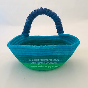 May include: A small, blue and green fabric basket with a blue fabric handle. The basket is woven in a circular pattern and has a slight oval shape. The text "© Leigh Hollmann 2020 All Rights Reserved www.swirlpuppy.com" is printed on the bottom of the basket.