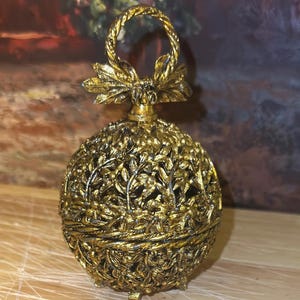 May include: A spherical gold-colored metal container with an intricate openwork leaf and vine pattern. The object features a hinged lid topped with a decorative bow and a circular handle, resting on small feet with a textured, antique finish.