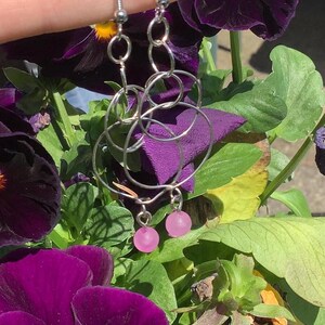 Può includere: Orecchini in filo argentato con un design unico e intrecciato. Ogni orecchino presenta una serie di cerchi interconnessi e una piccola perla rosa rotonda nella parte inferiore. Gli orecchini sono esposti su uno sfondo di vibranti fiori viola e fogliame verde.