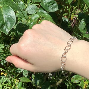 Könnte beinhalten: Ein silbernes Armband mit einer Reihe von miteinander verbundenen Kreisen. Das Armband wird an einem Handgelenk getragen, mit grünem Laub im Hintergrund. Ein filigranes Schmuckstück.