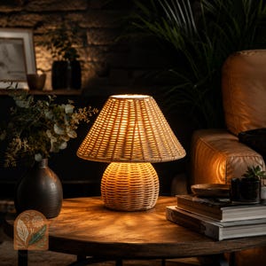 Peut inclure: Une lampe de table en rotin tressé avec un abat-jour conique et une base arrondie. La lampe est allumée, dégageant une lueur chaleureuse. Elle est posée sur une table ronde en bois, entourée d'objets décoratifs et de verdure.