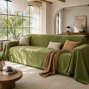 Puede incluir: Un sofá grande cubierto con una manta de color verde oliva texturizada con bordes de flecos. El sofá está decorado con tres cojines rectangulares en tonos crema y bronceado, y una manta bronceada doblada sobre el borde delantero.
