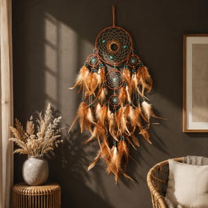 Puede incluir: Un atrapasueños grande con un aro central marrón con una red tejida, acentuado por cuentas turquesas y varios aros más pequeños. El diseño incluye numerosas plumas largas de color marrón claro que cuelgan de los aros, creando un efecto de capas.