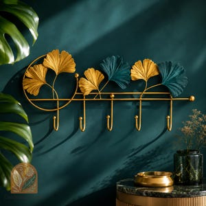 Peut inclure: Un porte-manteau mural en métal doré avec cinq crochets. Le porte-manteau présente des motifs de feuilles de ginkgo stylisées en or et sarcelle. Le porte-manteau est monté sur un mur bleu canard, avec un logo en bas à gauche.