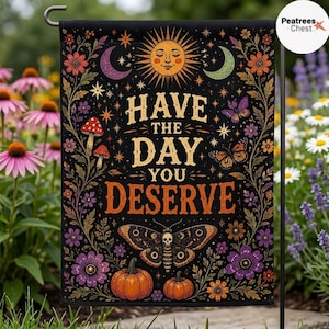 May include: A rectangular garden flag with a black background featuring a central sun, crescent moons, and the text HAVE THE DAY YOU DESERVE. The design includes colorful flowers, butterflies, a moth with a skull pattern, and two orange pumpkins.